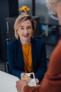 Smiling businesswoman holding POS system and colleague paying through credit card in office