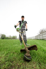 Professional gardener in a uniform is trimming the green grass. Male worker mows weeds with hand...