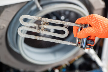 A hand in orange gloves holds the heating element of a washing machine after prolonged use in hard...