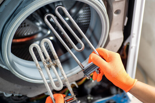 Hands in orange gloves hold two heating elements for a washing machine: one after a long time of use and the other new, against the background of a washing machine.