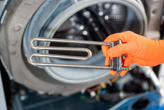 A hand in orange gloves holds a new heating element of a washing machine. Close-up against the background of a disassembled washing machine.