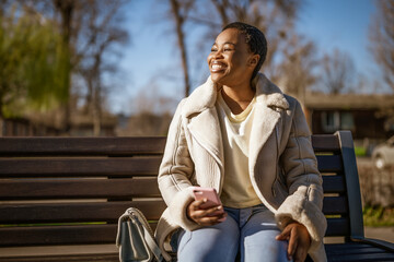 Outdoor portrait of happy african-american woman on sunny day. She is sitting on bench in the...