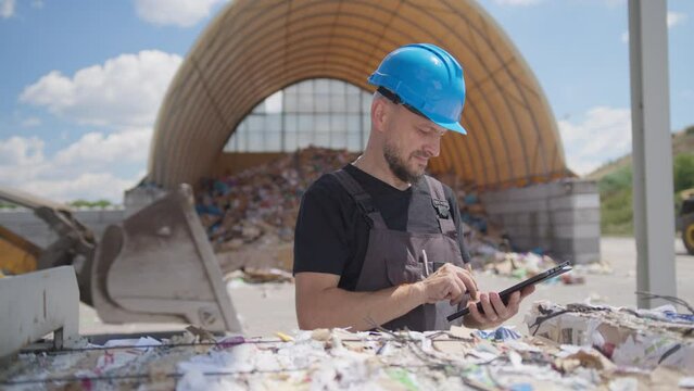 Male recycling plant worker uses tablet, tractor drives in background