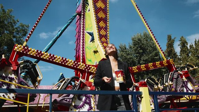 Shot of a positive, laughing charming woman holding a paper cup of popcorn and eating it with an amusement park in the background.