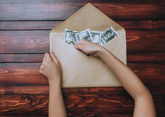 Two caucasian female hands hold an open beige craft envelope and take dollars on a brown wooden background. The concept of donation, alimony, winnings and cashback. Top view.