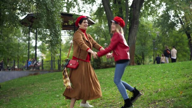 Wide shot joyful woman and girl spinning jumping on autumn park meadow holding hands. Cheerful relaxed Caucasian happy senior grandmother and teen granddaughter having fun on weekend outdoors