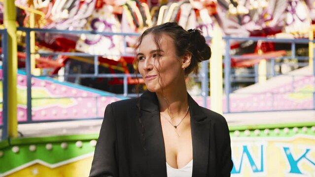 Shot of a positive, laughing charming woman holding a paper cup of popcorn and eating it with an amusement park in the background.