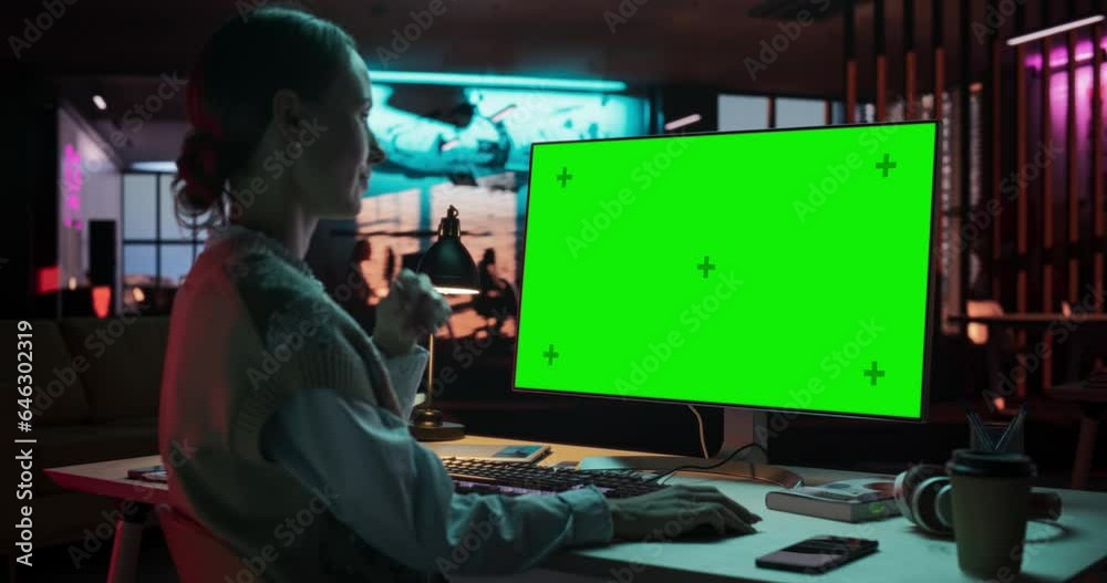 Poster over the shoulder: young woman sitting at her desk using desktop computer with mock-up green screen.