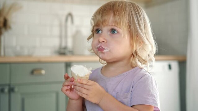 Baby girl enjoying ice cream. Pretty little toddler eating an ice-cream indoors, at home. Dining room background. Small child eats plombir and cream messy on her mouth. Cute kid with tasty sweet food.