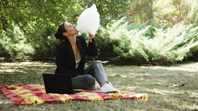 A business woman in a suit spends time outdoors in a city park on the grass, and eats cotton candy. Positive, carefree young woman rejoices after receiving a message on her laptop. Conclusion of a dea
