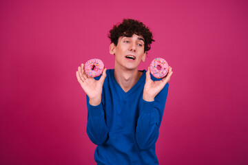 Young attractive curly man eating pink donuts and posing on a pink background.
