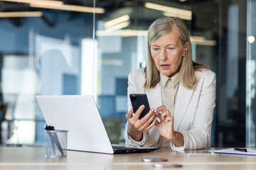 Mature adult business woman inside office at workplace received online notification message with bad news on phone, female boss with gray hair using phone frustrated and displeased reading online.