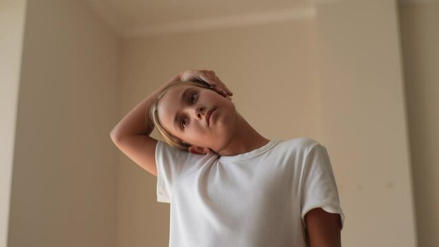 Close-up of serious little kid stretching neck for exercise during home workout standing in light living room. Pretty fitness girl child warm up. Concept of exercises for children at home.