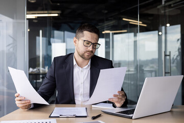 A worried young man businessman, accountant reviews documents, business agreements, bills in the office. He sits at the desk in the office and looks at the papers