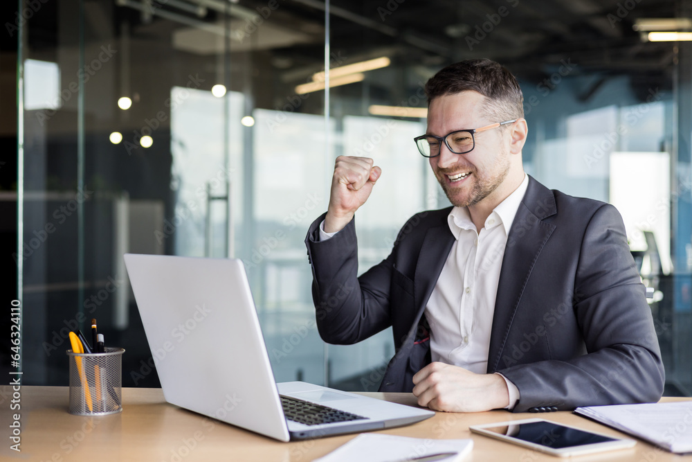 Wall mural Happy young man businessman rejoices success with hand gesture, looking at laptop screen, sitting in office at desk