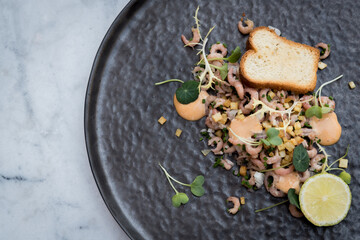 Close up of shrimp salad with toast and lime on black plate on a marble table