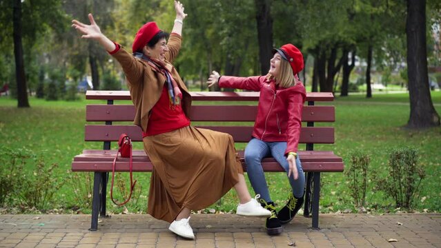 Wide shot cheerful senior grandmother and teen granddaughter stacking hands gesturing and hugging sitting on bench in park. Joyful carefree Caucasian woman and girl having fun enjoying weekend leisure