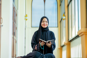 The image of an Asian Muslim woman in the Islamic religion in hijab in black  color. reading the Quran and having a happy Staying in a beautiful mosque, Arabic word translation: The Holy Al Quran.