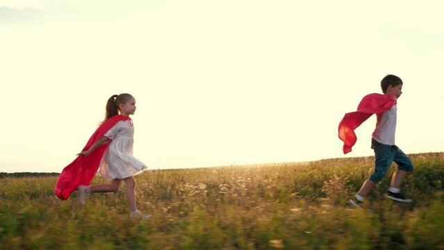 Joyful children group with red hero capes runs across rural field at back sunset