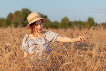 Naklejka na ściany i meble A young beautiful girl with braces on her teeth and long hair poses in a wheat field in the summer at sunset. The girl holds a hat in her hand against the background of a wheat field.