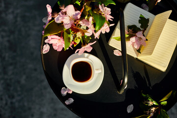Top view Cup of coffee flowers and notebook on the small black table on terrace at home in a sunny day, outdoor workspace, summer relaxation,  coffee break.