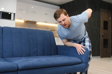 People, Healthcare and Treatment. Portrait of sick man suffering from acute side back pain, sitting on couch at home, person touching lower back aching area with hand. Health Problem Concept