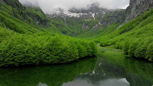 Spring mountain valley, lake on a foreground, clouds above mountain peaks, fly over the lake with reflection. Aerial opening shot 