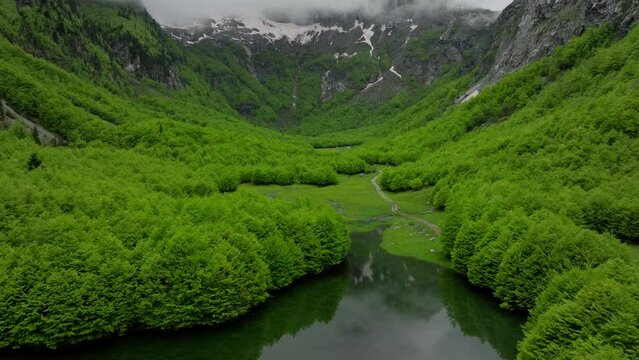 Green mountain valley in spring, clouds above mountains summit, fly over the lake with reflection. Aerial opening shot 
