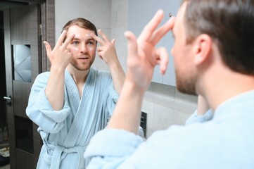 Handsome man looking at mirror and applying moisturizing cream on cheeks in bathroom. Groomed young guy doing skincare morning routine after taking a shower.