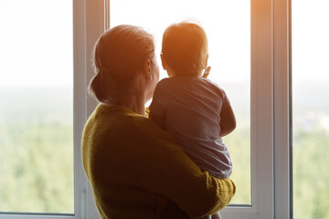 Grandmother holding a little cute baby girl and looks out the window at home, sunlight