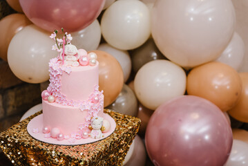 Birthday cake for 1 year on golden table. Pink cake is decorated with rabbit figure, unicorn and flowers decor for girl. Delicious reception at party. Trendy cake on background of balloons. Closeup