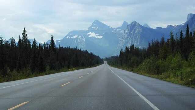 Driving down the icefields parkway in the Rocky Mountains, Alberta, Canada. Road through tall snowcapped mountains and green pine forests. 
