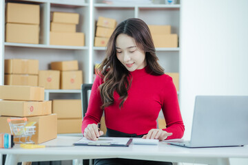 Start a small business Young independent Asian woman using laptop computer working with boxes in home office Young Asian woman smiling happy at successful sales Packaging boxes, online marketing and s