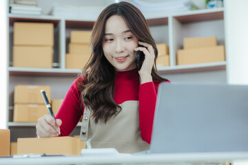Start a small business Young independent Asian woman using laptop computer working with boxes in home office Young Asian woman smiling happy at successful sales Packaging boxes, online marketing and s