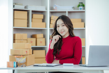 Obraz na płótnie Canvas Asian woman small business owner starting an SME. Independent Asian woman using laptop working with boxes at home. Young Asian woman who succeeds by raising her hand Packaging boxes, online marketing 