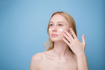 A beautiful girl applies moisturizer to her facial skin. Young woman on blue background smiling and...