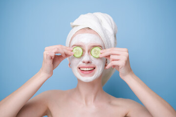 Young woman with face mask and towel on her head holding fresh cucumber slices. Happy girl taking care of her facial skin on blue background.
