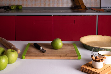 One green apple on a cutting board and kitchen knife on a table with ingredients for apple pie: green apples, cinnamon sticks and raw crust pie dough. Cooking homemade Thanksgiving fruit autumn tart