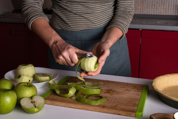 Woman is peeling apples to prepare apple pie in the kitchen. Authentic female hands peel an apple with a knife. Cooking apple pie process, recipe step by step. Thanksgiving preparation, autumn bakery