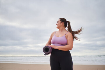 Body positive lady smiling in the ocean beach with a yoga mat and looking at the water