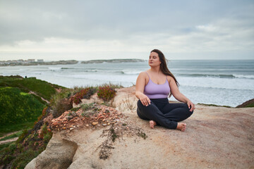 Fototapeta na wymiar Full figured woman looking away and meditating while sitting at gloomy wild beach