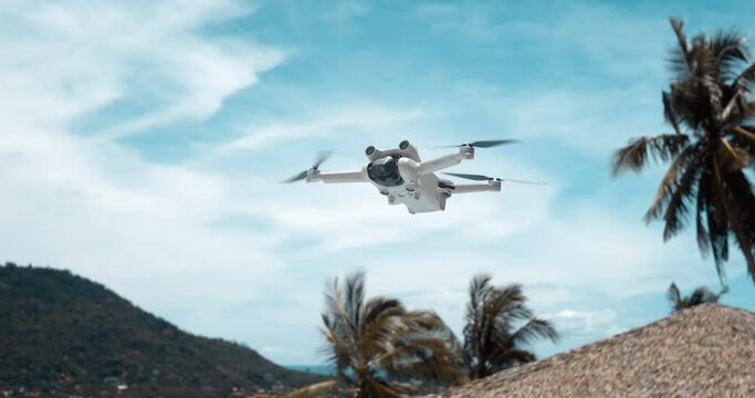 Drone aircraft with blurred fast rotating propellers and video camera flying in air in slow motion. Drone copter flying in air on blue sky and palms background. Making content while summer traveling