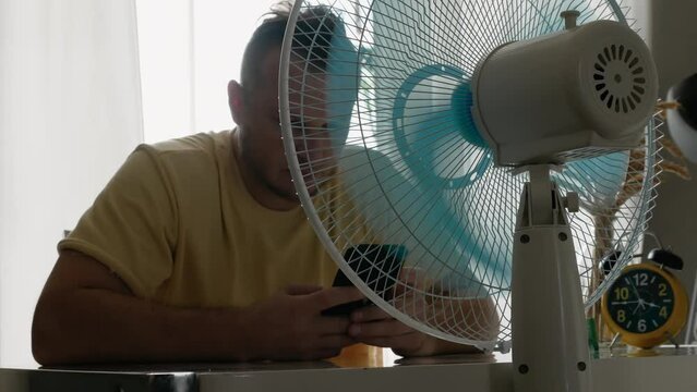 An electric fan cools the air. The person is very hot, the fan is the air duct in the apartment. Man cools down with a smartphone in his hands