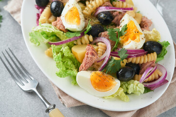 Tuna salad with pasta, eggs, potatoes, olives, red onions and sauce in white plate on old light gray concrete table background. Nicoise salad. French cuisine. Top view. Flat lay.