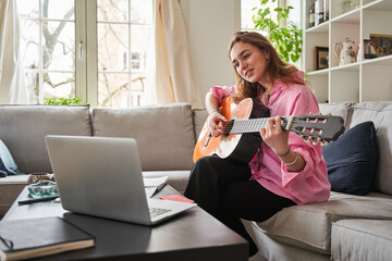 Teen girl learning to play guitar at home while watching online laptop lessons