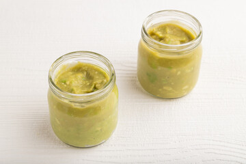 Baby puree with vegetable mix, broccoli, avocado in glass jar on white wooden, side view