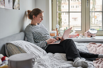 Calm teen woman wearing domestic clothes sitting on the bed and looking at laptop - 646387594