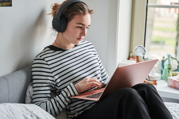 Young teen woman wearing headphones sitting on bed and looking at laptop - 646387727