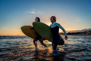 Two surfers with surfboard prepares to hit the waves at sunset.