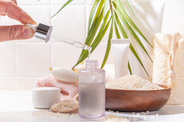 Rice Water Serum. Natural organic rice cosmetics. Fermented beauty care, wellness and spa. Transparent white serum drops bottle with rice grain and bath utensils, on white bathroom background